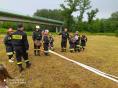 Sześcioro dzieci w mundurach strażackich i trzech dorosłych strażaków OSP w mundurach rozwijają węże strażackie na wykoszonym polu. W tle kładka nad rzeką 