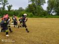 Grupa dzieci i strażaków OSP w mundurach biegnie z wężami strażackimi po wykoszonym polu. W tle zielone drzewa
