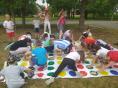 Grupa dzieci w letnich ubraniach gra w grę twister na położonej na trawie dużej białej planszy z szeregami kolorowych kół. W tle dom, droga i zielone drzewa