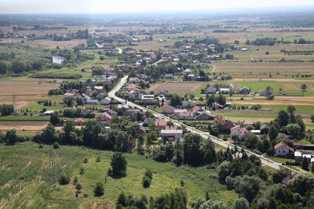 Widok wsi z lotu ptaka. Wzdłuż drogi posadowione domy, zielone drzewa i zielone i żółte pola. Po lewej stronie zakręt rzeki porośnięty krzewami